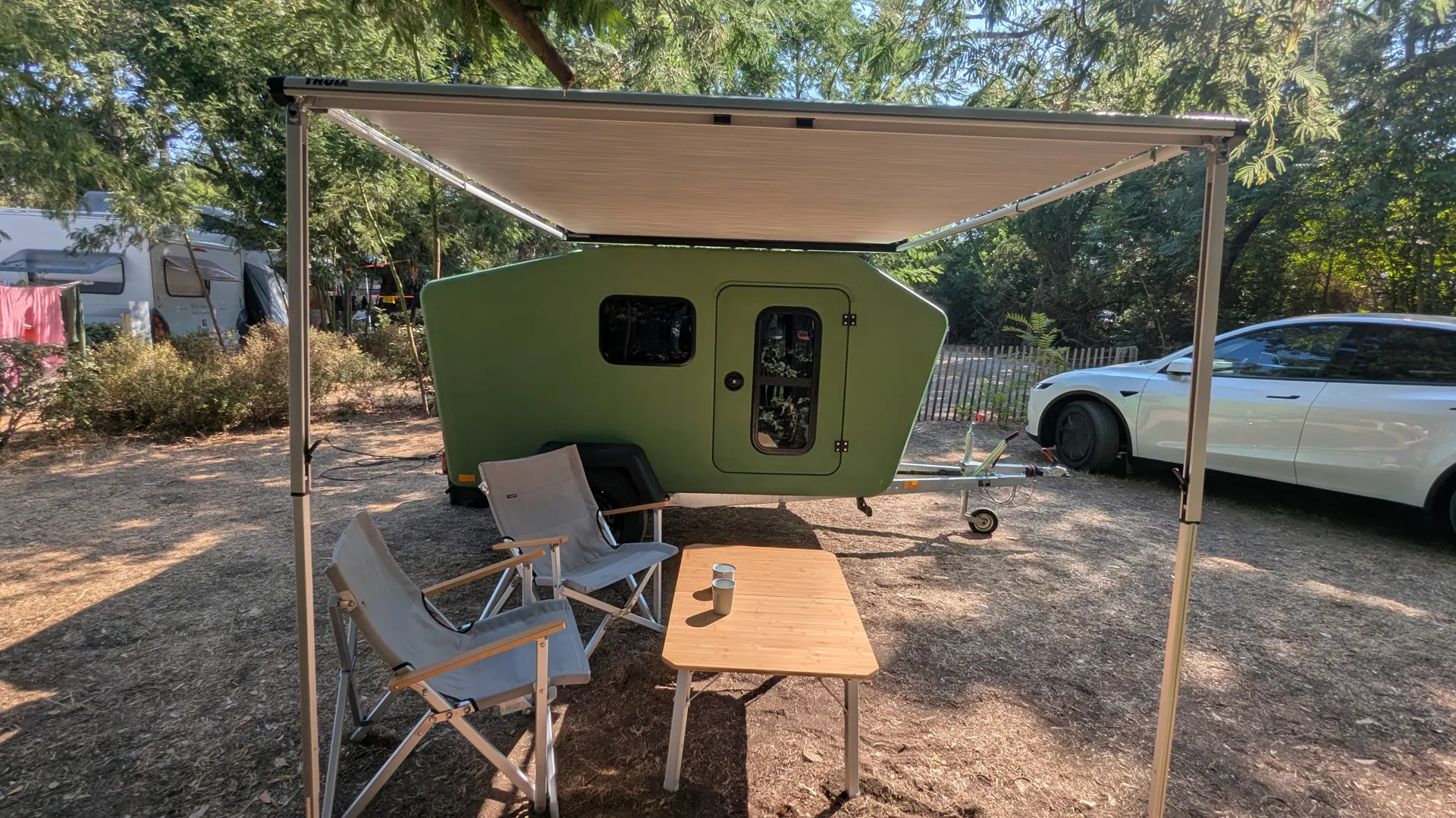 Extended awning over two camping chairs and a bamboo table in front of the green Glide at a campsite